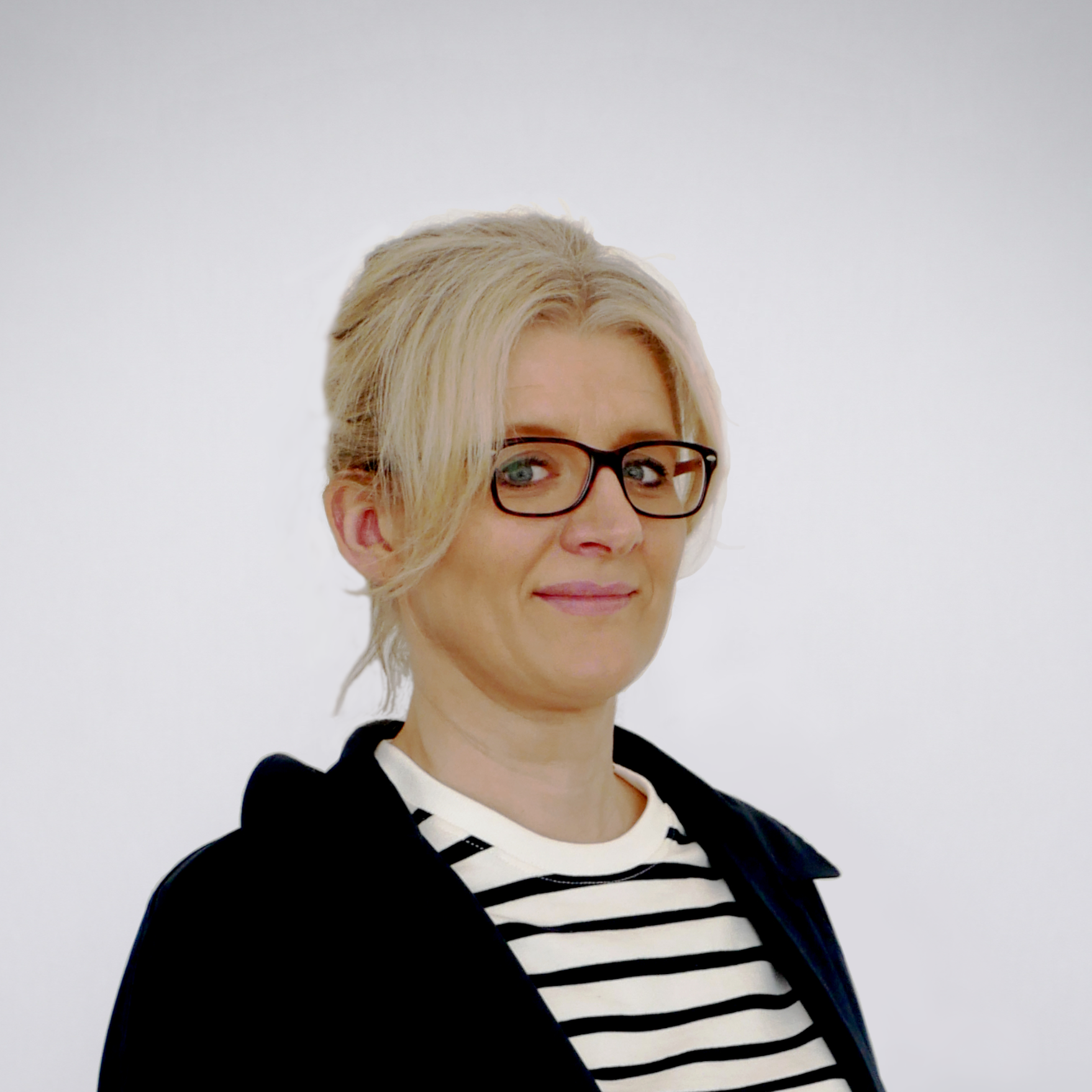 Auf diesem Bild ist Frau Frenger ab Mitte des Brustkorbes abgebildet. Sie steht leicht nach links gedreht, schaut frontal zur Kamera und lächelt. Sie hält die Arme jeweils seitlich am Körper. Frau Frenger trägt einen offenen, dunkelblauen Blazer über einem weiß-blau-quergestreiften Oberteil mit rundem Ausschnitt. Frau Frenger trägt ihr langes, blondes, Haar zu einem Zopf gebunden und hochgesteckt mit einzelnen Strähnen im Stirnbereich, die seitliche am Gesicht herabhängen. Sie trägt eine Brille mit dunklem Gestell und hat graublaue Augen. Der Hintergrund des Fotos ist seicht blaugrau gefärbt mit einer zur Mitte hin zunehmenden Aufhellung.