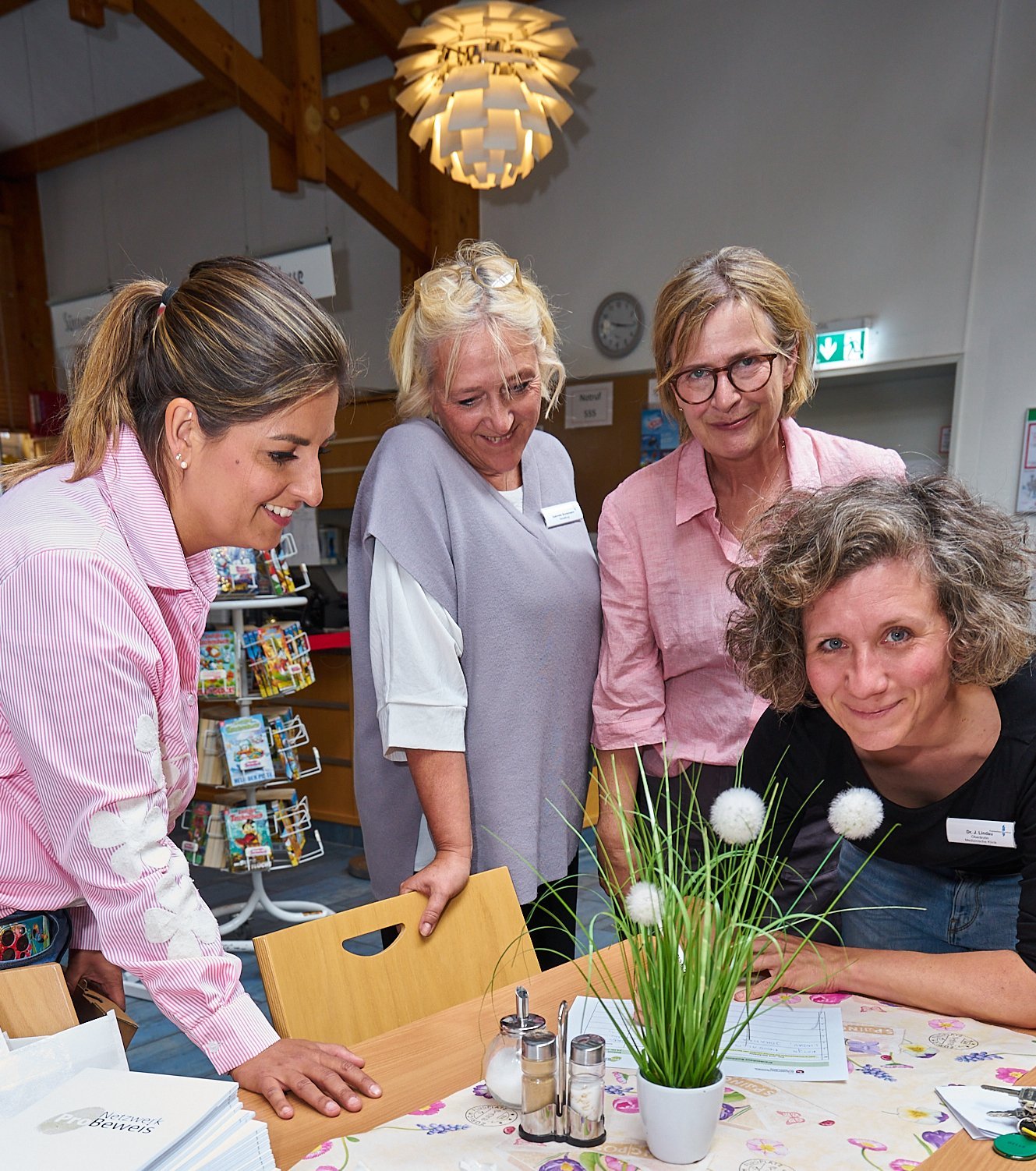 Zu sehen sie Frau Stockhausen sowie drei Mitarbeitende des Krankenhauses Buchholz. Sie stehen um einen Tisch versammelt, wobei sich ein Mitarbeitende auf den Tisch auflehnt, eine mit der Hand auf einer Stuhllehne und Frau Stockhausen sich mit einer Hand auf dem Tisch aufstützt. Augenscheinlich wird ein Dokument, welches auf der Tischfläche liegt angesehen / besprochen. 
