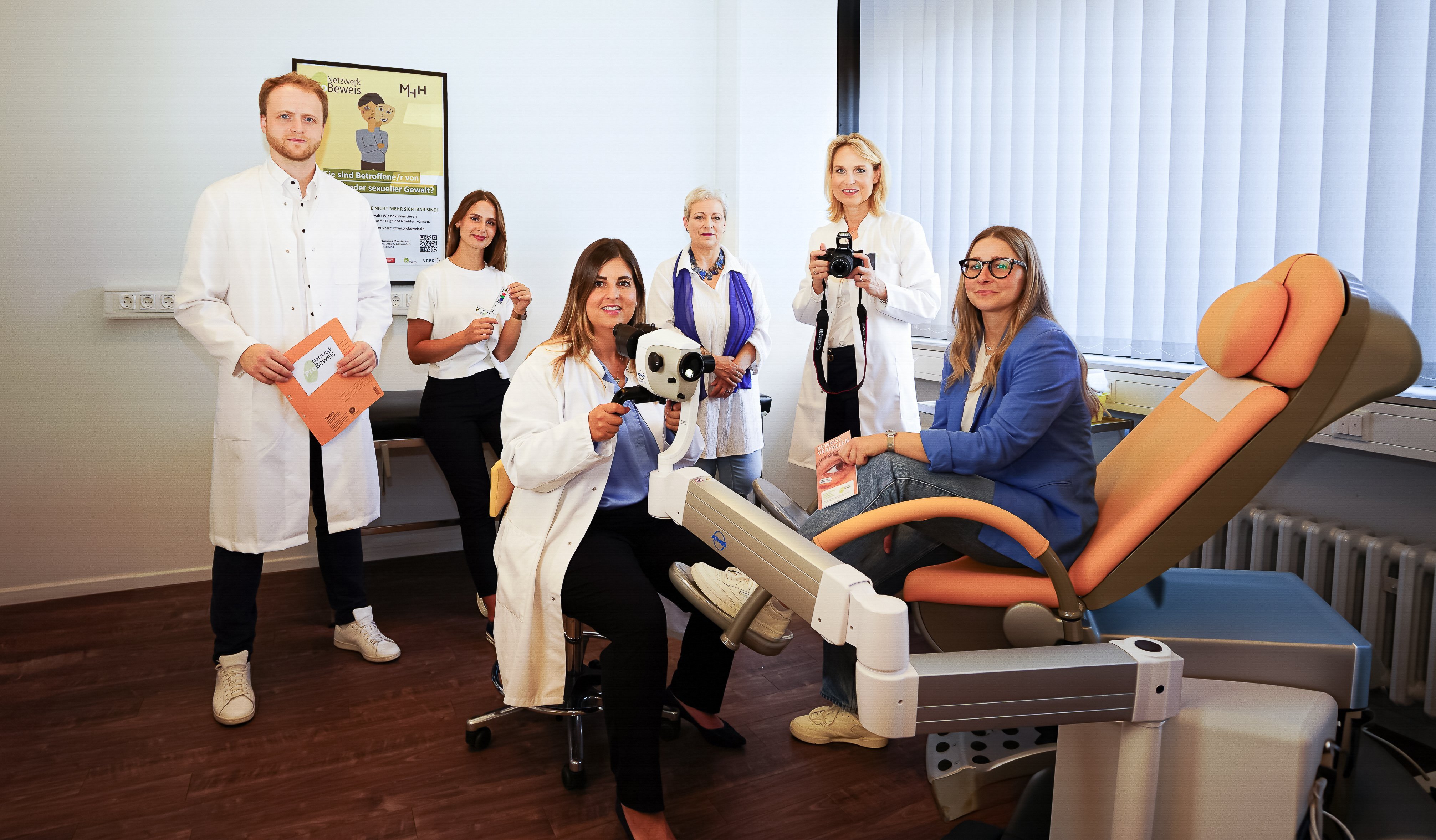 Das Team vom Netzwerk ProBeweis im Untersuchungsraum des Instituts für Rechtsmedizin. Im rechten Vordergrund befindet sich ein gynäkologischer Untersuchungsstuhl mit gelbem Bezug. Darauf sitzt in entspannter Haltung mit nur einem Bein auf der dafür vorgesehenen Fußstütze aufgestützt, sowie mit einem weißen T-Shirt, einem blauen Sakko, einer Jeans und weißen Schuhen bekleidet, eine junge, freundlich in die Kamera lächelnde Frau mit langen, blonden Haaren und einer Brille. Sie hält in ihrer linken Hand einen Flyer vom Netzwerk ProBeweis, welche auf dem linken, aufgestützten Knie aufliegt. Vor ihr, weiter links auf dem Bildausschnitt, sitzt eine weitere, offen Richtung Kamera lächelnde Frau mit langen, glatten, braunen Haaren und hellen Strähnen auf einem hellgelb gepolsterten Drehstuhl vor einem, zum gynäkologischen Stuhl gerichteten, sogenannten Kolposkop. Ein Kolposkop ist eine Apparatur mit einer Kombination aus Lichtquelle, Mikroskop mit Okular und Fotoapparat. Dieses Kolposkop hält die Frau mit beiden Händen an den dafür vorgesehenen Griffen fest. Sie ist mit einer blauen Bluse und einer schwarzen Hose sowie einem weißen Arztkittel bekleidet. Etwas weiter in der Tiefe des Raumes, hinter dem gynäkologischen Stuhl, steht eine weitere, freundlich in die Kamera lächelnde Frau mit schulterlangen blonden Haaren, einer weißen Bluse, einer schwarzen Hose und ebenfalls einem weißen Arztkittel. Sie hält in beiden Händen, angehoben vor dem Körper, eine einsatzbereite, professionelle Digitalkamera. Zu ihrer rechten und somit weiter links auf dem Bildausschnitt, etwa hinter dem Kolposkop in der Tiefe des Raumes, steht eine dezent lächelnde und frontal zur Kamera stehende, ältere Dame mit kürzeren, weißgrauen Haaren, einer weißen Bluse, einem übergeworfenen, blauen Seidentuch und einer hellblauen Jeans. Sie steht an eine, hinter ihr an der Wand stehende Untersuchungsliege leicht angelehnt und hält die Hände vor dem Bauch verschränkt. Hinter der rechten Schulter in einem lockeren Abstand steht eine weitere junge Frau an die Untersuchungsliege gelehnt beziehungsweise halb auf ihr sitzend. Sie trägt lange, offene, braune Haare, ein weißes T-Shirt sowie eine schwarze Hose. Sie steht ebenfalls nahezu frontal zur Kamera und lächelt freundlich. Mit beiden Händen hält sie einen forensischen Untersuchungswinkel mit Maßstab und Farbskala vor sich. Rechts daneben sowie links am Bildrand steht ein Mann mit weißem Hemd, schwarzer Hose, weißen Schuhen, mit mittelblonder Kurzhaarfrisur und Bart. Er lächelt dezent in die Kamera und hält mit beiden Händen eine orangefarbene Mappe mit der Aufschrift und dem Fingerabdruck-Logo vom Netzwerk ProBeweis vor dem Körper. Über der Untersuchungsliege an der Rückwand des Bildausschnittes hängt ein Bilderrahmen mit dem Poster vom Netzwerk ProBeweis. Links hinter dem gynäkologischen Stuhl befindet sich eine große Fensterfront mit Jalousien, durch die Tageslicht in den Raum strömt.