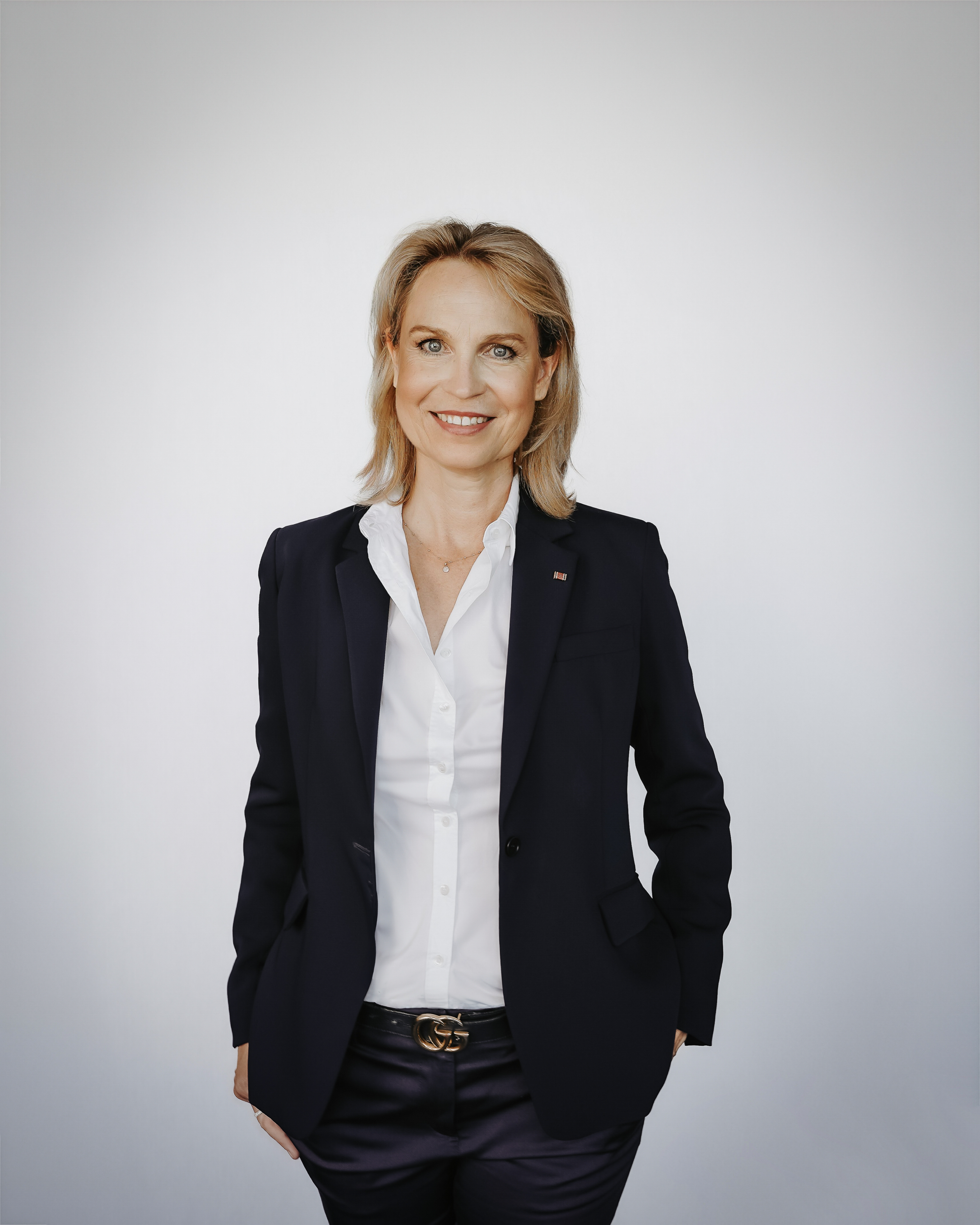 Auf diesem Bild ist Professorin Debertin ab dem oberen Drittel der Oberschenkel abgebildet. Sie steht frontal zur Kamera und hält die Hände beidseits eng an den Hüften, was teilweise von ihrem Blazer verdeckt wird. Sie trägt eine weiße Bluse unter einem dunkelblauen Sakko zu einer dunkelblauen Hose. An der äußeren Kante des linken Revers ist eine rot-schwarz-goldene Ordensminiatur angesteckt. Um die Taille in den Hosenschlaufen befindet sich ein dunkelblauer Gürtel mit einer goldfarbenen Schnalle. Um den Hals trägt sie eine kurze, goldfarbene Kette mit kleinem runden Anhänger. Professorin Debertin lächelt freundlich mit einem halboffenen Mund, sodass die obere Zahnreihe sichtbar ist. Sie trägt blonde Haare über Kinnlänge mit einem rechtsseitigen Scheitel und hat blau-grüne Augen. Der Hintergrund des Fotos ist seicht blaugrau gefärbt mit einer zur Bildmitte hin zunehmenden Aufhellung.