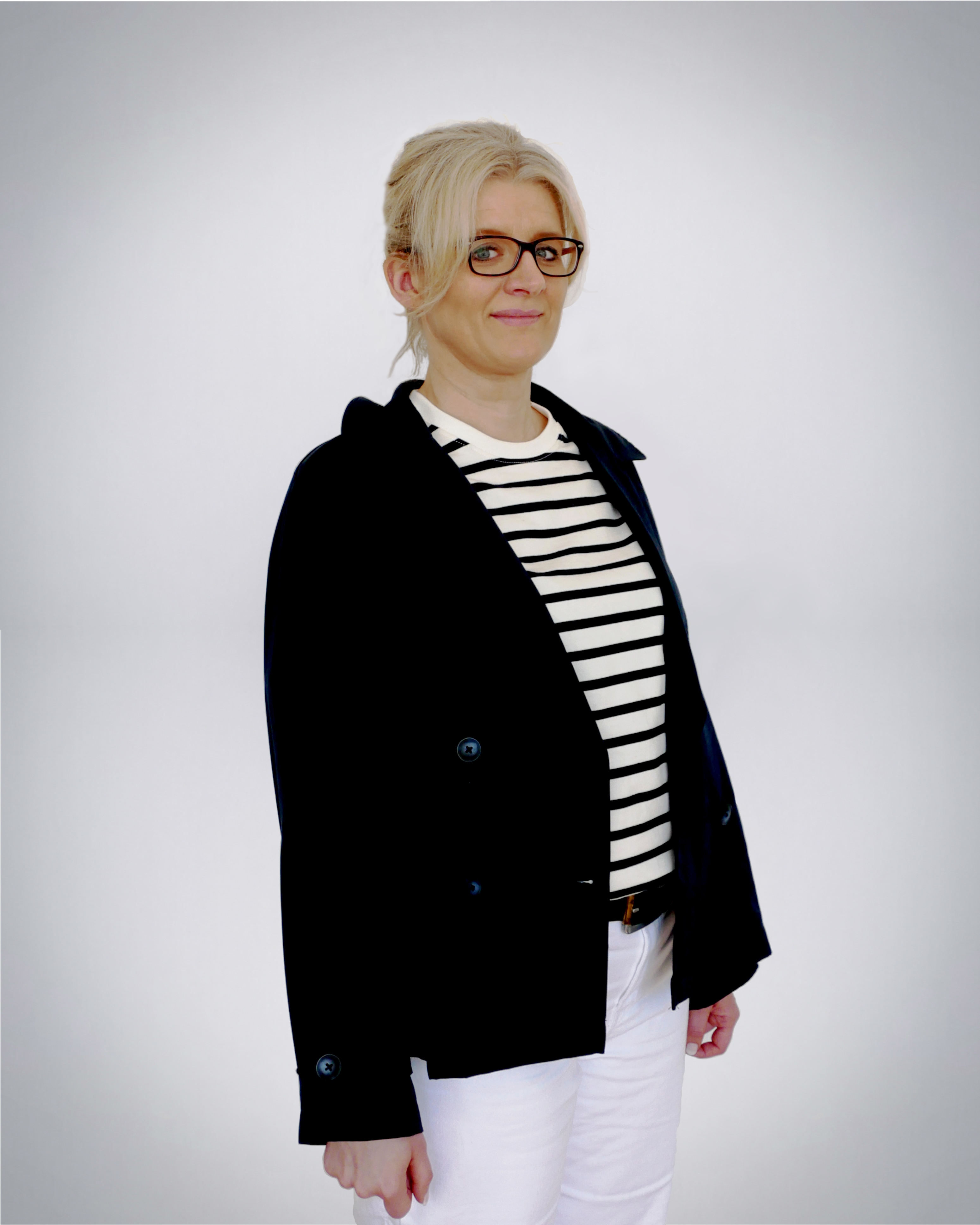 Auf diesem Bild ist Frau Frenger ab Mitte der Oberschenkel abgebildet. Sie steht leicht nach links gedreht und schaut frontal zur Kamera und lächelt. Sie hält die Arme jeweils seitlich am Körper. Frau Frenger trägt einen offenen, dunkelblauen Blazer über einem weiß-blau-quergestreiften Oberteil mit rundem Ausschnitt sowie dazu eine weiße Hose mit dunklem Lebergürtel. Frau Frenger trägt ihr langes, blondes, Haar zu einem Zopf gebunden und hochgesteckt mit einzelnen Strähnen im Stirnbereich, die seitliche am Gesicht herabhängen. Sie trägt eine Brille mit dunklem Gestell und hat graublaue Augen. Der Hintergrund des Fotos ist seicht blaugrau gefärbt mit einer zur Mitte hin zunehmenden Aufhellung.