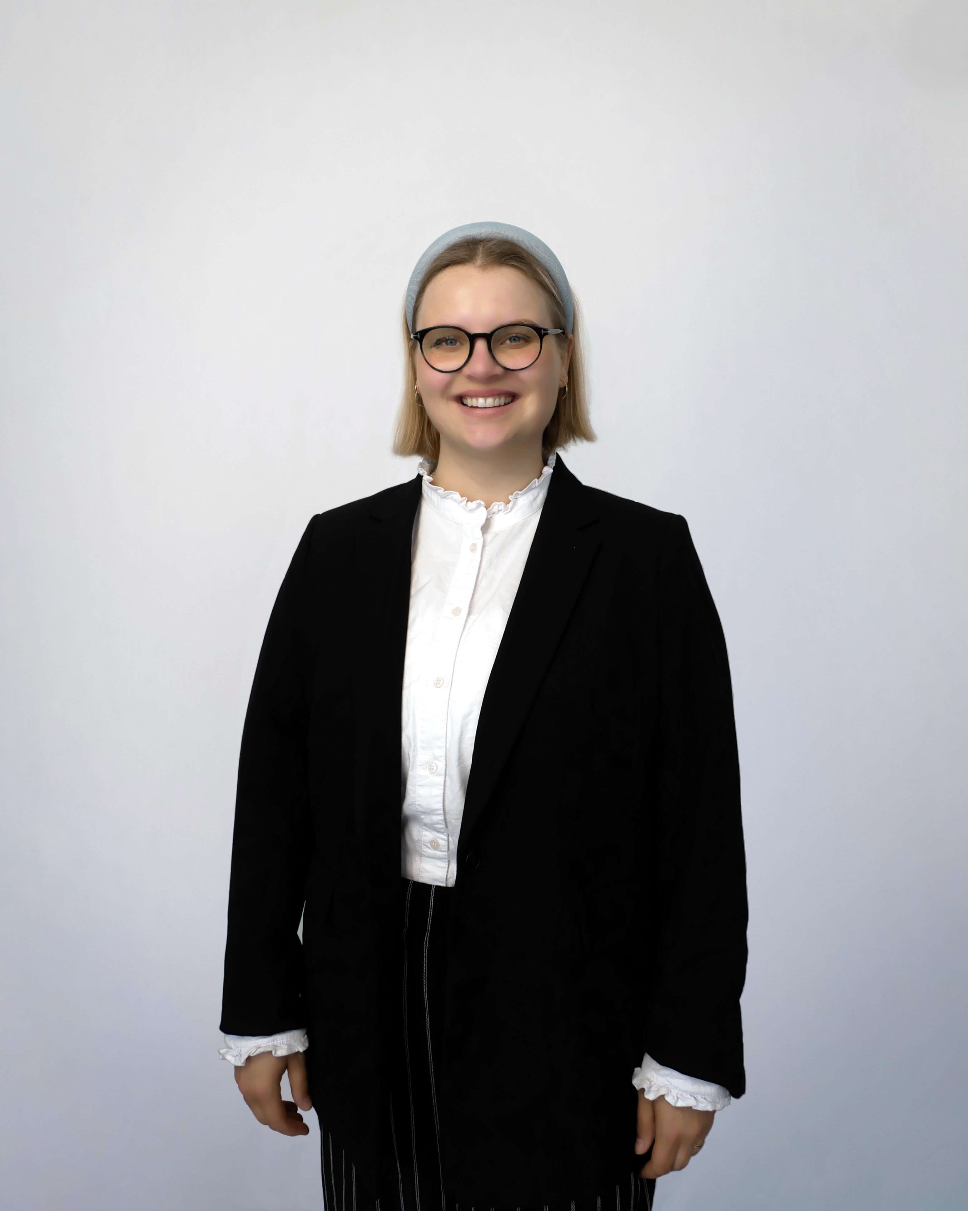 Auf diesem Bild ist Frau Krüger ab etwa der Hälfte der Oberschenkel abgebildet. Sie steht etwas leicht nach rechts gedreht nahezu frontal zur Kamera. Ihre Arme hängen seitlich herab. Sie trägt eine weiße Bluse unter einem schwarzen Sakko zu einer schwarzen, mit weißen Nadelstreifen versehenen Hose. Sie trägt goldfarbene Kreolen sowie eine Brille mit runden Gläsern und schwarzem Gestell. Ihre Augenfarbe ist blau. Frau Krüger lächelt freundlich mit halboffenem Mund, sodass die obere Zahnreihe sichtbar ist. Sie trägt blonde Haare bis etwa zur Höhe des Kehlkopfes und einen hellblauen Haarreif. Der Hintergrund des Fotos ist seicht blaugrau gefärbt mit einer zur Bildmitte hin zunehmenden Aufhellung.