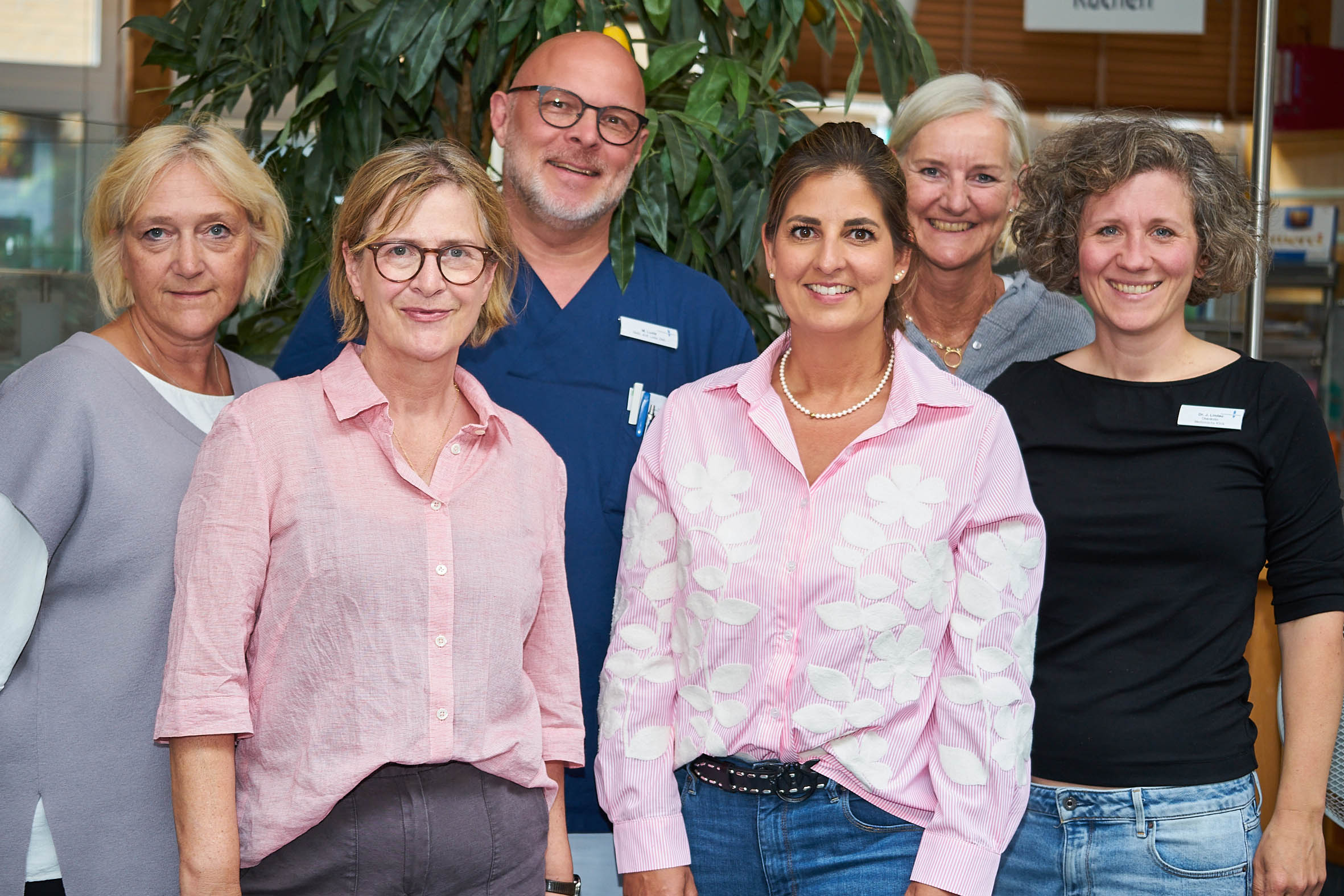 Zu sehen sind sechs Personen in zwei voreinander stehenden Dreierreihen in einer halbtotalen Aufnahme. Frau Stockhausen steht in der vorderen Reihe in der Mitte. Die übrigen Personen repräsentieren das Klinikpersonal des Krankenhaus Buchholz. Alle Personen schauen freundlich in die Kamera und haben ihre Arme, soweit dargestellt, locker der Seite anliegend herabhängen. 