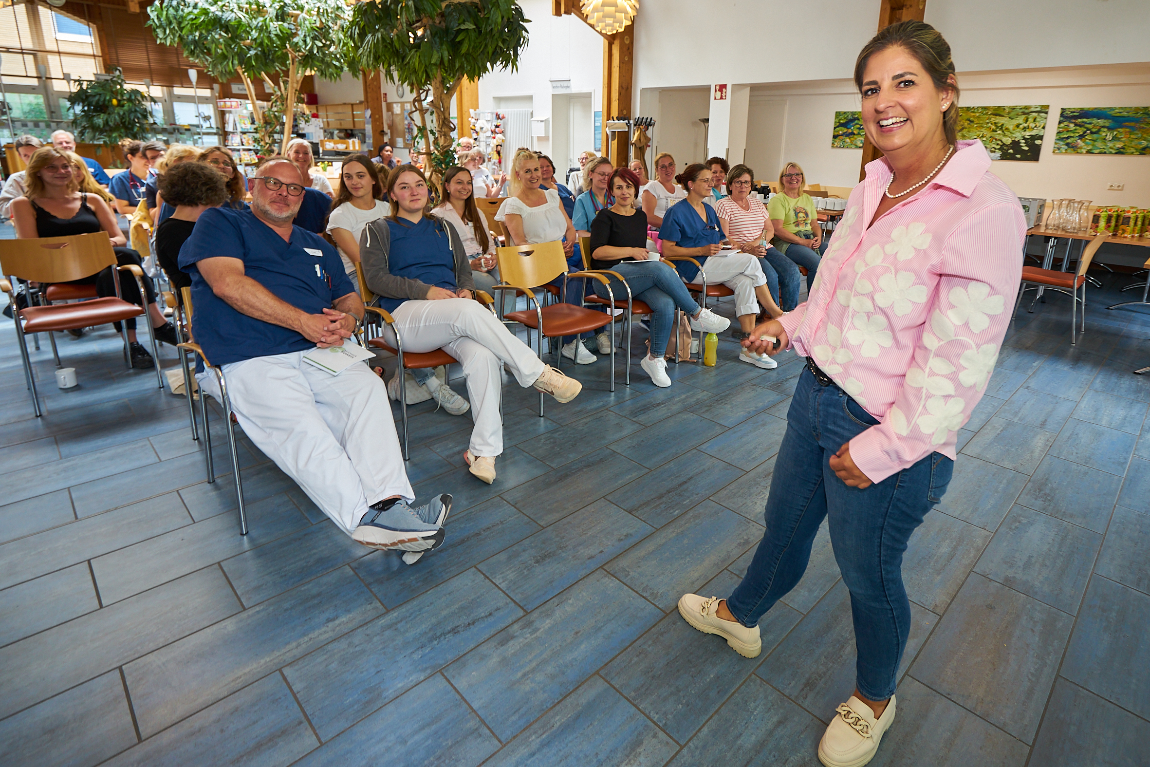 Zu sehen ist, wie Frau Stockhause in einem größeren Raum vor einer in Stuhlreihen sitzenden Menschenmenge von etwa dreißig Personen steht und freundlich in die Kamera lächelt. In der rechten Hand hält sie einen Presenter und scheint gerade ihren Vortrag zu halten. In der Zuhörerschaft scheinen die überwiegend in typischer Krankenhaus-Arbeitskleidung gekleideten Anwesenden ihrem Vortrag freundlich und fröhlich zu folgen. 